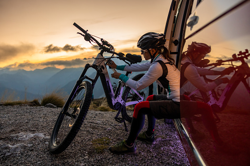 Female cyclist with long brown hair tied in a ponytail,wearing helmet and sports clothing plugging in a cable to charge her e-bike on top of a mountain while sitting in a red van,side view during sunset