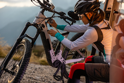 Adult woman with long brown hair tied in a ponytail,wearing helmet and sports clothing plugging in a cable to charge her e-bike during a break from her biking trip to the top of Mangart