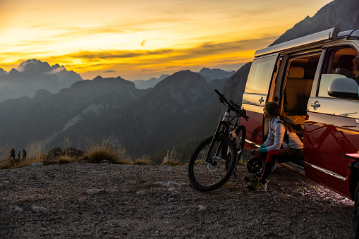 Adult woman with long brown hair tied in a ponytail taking a break from her biking trip,sitting inside a red van with opened door and looking at the beautiful view,bike standing in front of her during sunset,wide angle shot