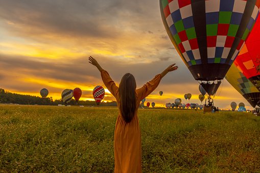 Woman with long brown hair wearing a orange colored dress,lifting her arms while looking at hot air balloons taking off on a agricultural field