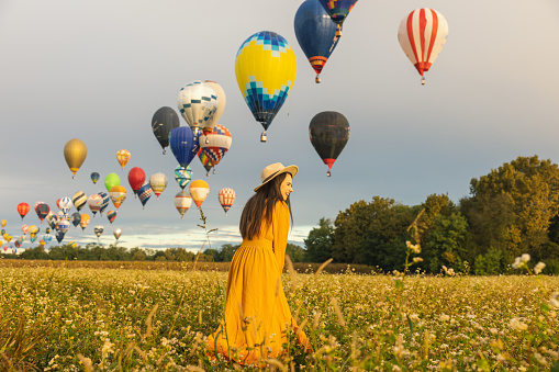 Woman with long brown hair,wearing a orange colored dress,laughing while standing in a flower field with lots of different colored hot air balloons in the background