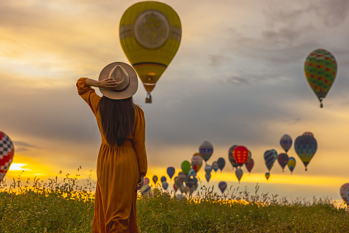 Woman with long brown hair wearing a orange colored dress,and straw hat looking at hot air balloons flying in the air over meadow during sunrise,holding her straw hat