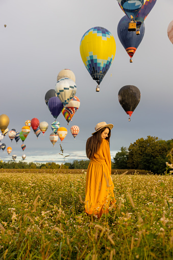 Woman with long brown hair,wearing a orange dress,smiling while standing on a flower meadow during sunrise,plenty of colorful hot air balloons in background