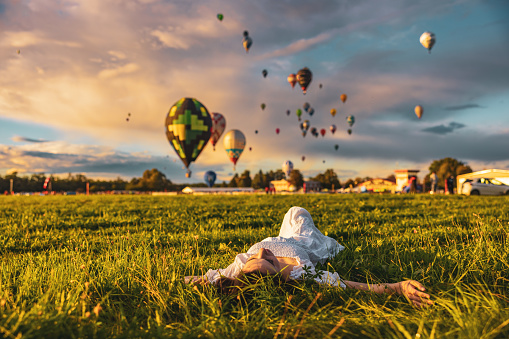 Woman with long brown hair,wearing a white dress,lying down on meadow,looking in the sky,hot air balloons in background