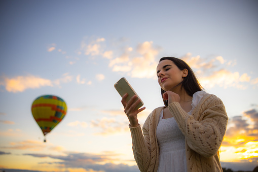 Woman with long brown hair,wearing a white dress and beige pullover,surfing the net on her smartphone while a beautiful hot air balloon is soaring in the sky behind her,early morning