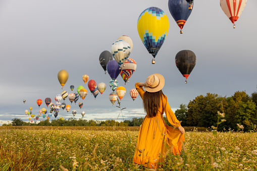 Woman with long brown hair wearing a orange colored dress,holding her straw hat while looking at hot air balloons flying in the air over agricultural field during sunrise