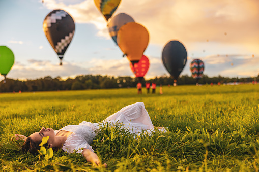 Woman with long brown hair,wearing a white dress,and closed eyes lying down with stretched out arms on a meadow,hot air balloons in background