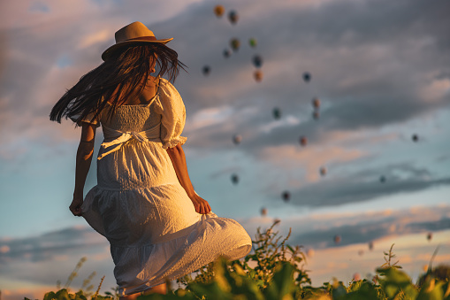 Woman with long brown hair,wearing a white dress while walking on a meadow,holding her dress,lots of colorful hot air balloons in the background,rear view
