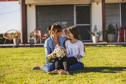 Mother with long brown hair and daughter with long brown hair smiling at each other while kneeling on the lawn in the backyard of their house,holding daisy flowers