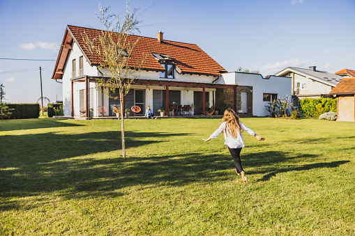 Little girl with long brown hair is running through the garden on lawn with a tree next to her,spreading her arms while running,house in the background,during summertime,rear view