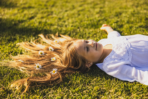 Girl with long brown hair is smiling while lying down on a lawn with outstretched arms and closed eyes with flowers in her hair,side view during summertime and daylight