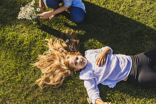 Close-up girl with light brown hair is lying down on the lawn in the backyard of her house,has flowers in her hair,mother is kneeling next to her and putting the flowers in her hair,daughter smiling and looking at camera,summertime,high angle view