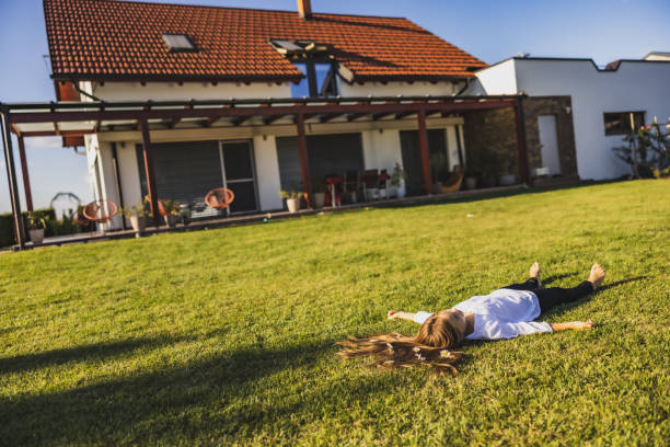 petite fille légère allongée sur la pelouse dans l’arrière-cour de sa maison avec les bras tendus, regardant dans le ciel - child blank expression pensive focus on foreground photos et images de collection