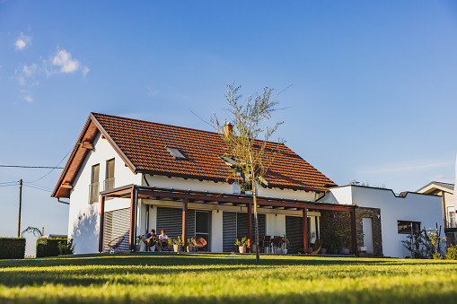 Modern house with terrace on the countryside,two boys sitting on chairs on the terrace and using their phone,large backyard during summertime,young tree in front,clear sky in the background,low angle view