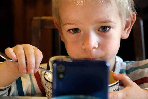 garotinho usando telefone enquanto come sopa durante o almoço ou jantar na mesa. criança comendo comida e assistir desenho animado no smartphone porque nomofobia ou transtorno de déficit de atenção com hiperatividade tdah - little boys breakfast caucasian child - fotografias e filmes do acervo