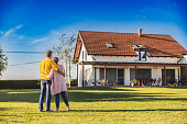 Couple,man and woman,hugging each other while standing on the lawn in the backyard of their new bought house,rear view,modern house in the background