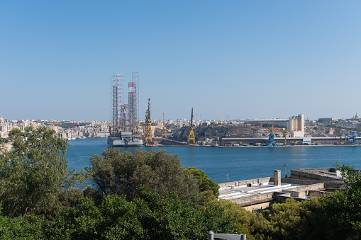 Magnificent view of the sea bay and buildings of the old city from the fortress wall, medieval Maltese architecture, forts and palaces, cathedrals and residential buildings, classical style, cityscape, ships in the harbor, port cranes, sunny day, summer, Europe.