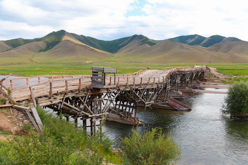 Old bent and deformed wooden bridge over river against mountain range, Jargalant, Mongolia.