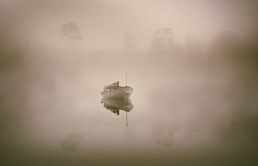 A boat sits on the still waters of a misty lake. Through the fog we can see the tree-lined shore.