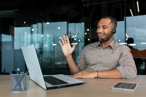 Online training. A young African-American male student sits in an office at a desk in front of a laptop in a headset. Learns through a video call, says hello, smiles.
