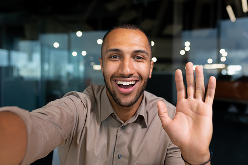 Smiling and cheerful businessman in office taking selfie photo on phone and talking on video call with colleagues and friends using smartphone, african american man waving at camera greeting gesture