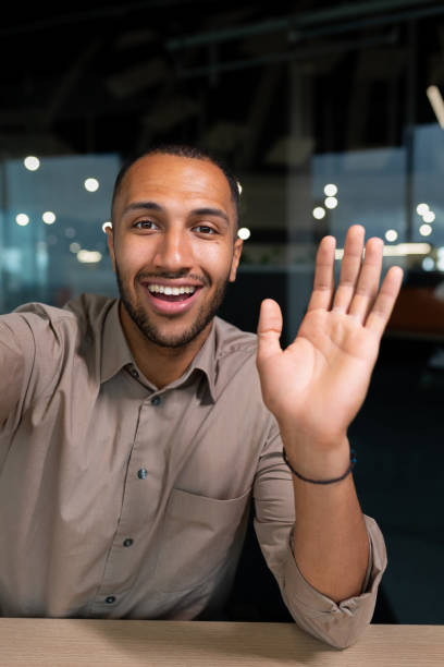 lächelnder und fröhlicher geschäftsmann im büro, der selfie-fotos am telefon macht und per smartphone mit kollegen und freunden per videoanruf spricht, afroamerikanischer mann winkt in die kamera-begrüßungsgeste - photographing smart phone friendship photo messaging stock-fotos und bilder