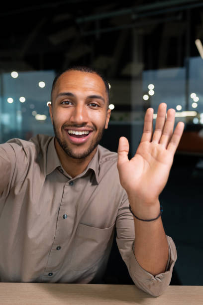 smiling and cheerful businessman in office taking selfie photo on phone and talking on video call with colleagues and friends using smartphone, african american man waving at camera greeting gesture - filming point of view fotos imagens e fotografias de stock