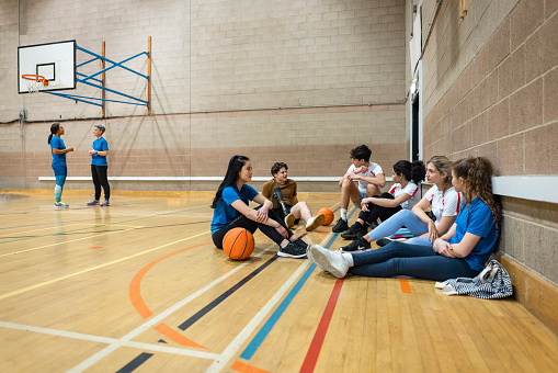 A wide-angle shot of a diverse group of teenagers sitting on the floor during the halftime of a basketball game. They are chatting and socialising amongst themselves.
