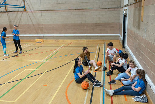 A wide-angle shot of a diverse group of teenagers sitting on the floor during the half time of a basket ball game. They are chatting and socialising amongst themselves.