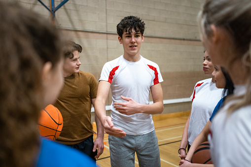 An over-the-shoulder shot of a group of secondary school students both male and female discussing and sharing feedback with each other regarding the basket ball game. They are all listening and maintaining eye contact with each other.