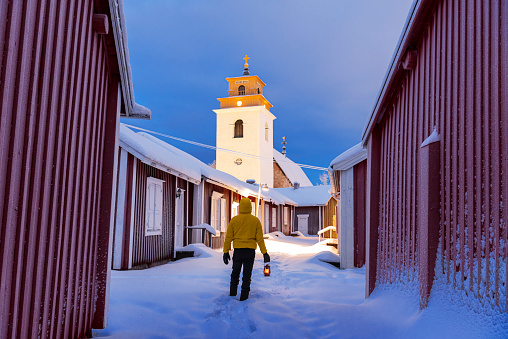 Winter view of a man with lantern in the snowy street of the illuminated medieval town of Gammelstad at dusk, UNESCO world heritage site, Lulea, Norrbotten, Sweden, Europe