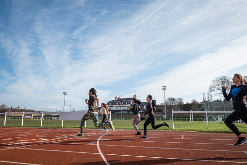 A side shot of a diverse group of females training on a running track. It is a bright day with blue skies. There are no spectators in the frame.