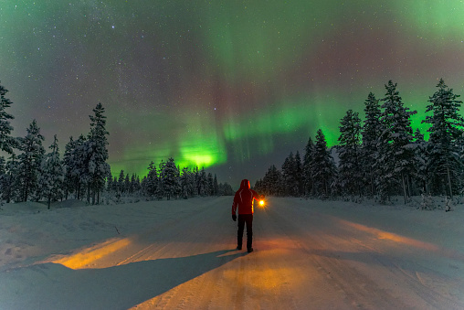 Man in red jacket standing in the middle of a snowy road with lantern in his hand watching northern lights, Lapland, Sweden.