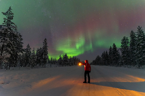 View of a man with lantern standing in the middle of the road going through the forest looking at northern lights (aurora borealis), Lapland, Sweden