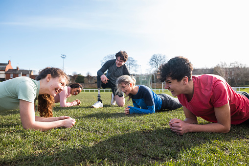 A shot of a group of teeneagers holding a plank in the press up possiton on an outdoor sporintg field. They are smiling as they compete with each other.