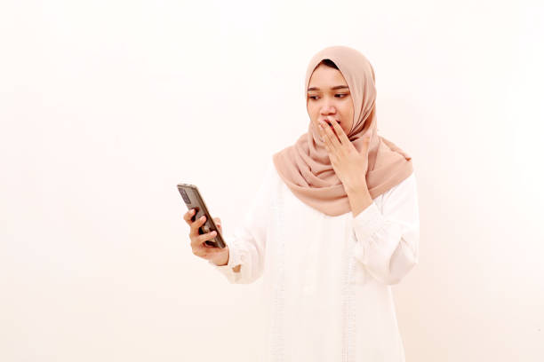 Young asian muslim woman holding a cellular phone with shocked face expression. Isolated on white Young asian muslim woman holding a cellular phone with shocked face expression. Isolated on white no anger and negativity in ramadan stock pictures, royalty-free photos & images