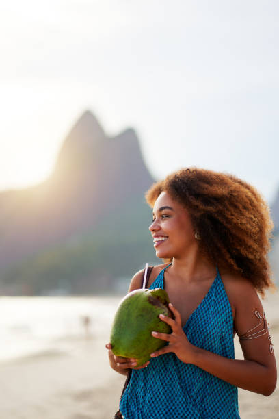 ブラジルの黒人女性は、イパネマの日没時にドスエルマノス山を背景にビーチでココナッツを手に持つ - nature curly hair smiling human face ストックフォトと画像