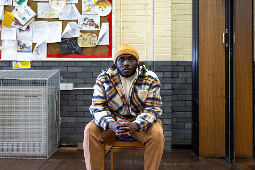 A full shot of a young adult sitting, leaning forward in casual clothing looking at the camera in a community centre. He has a negative emotion on his face. The community centre is located in Seaton Deleval in the North East of England.