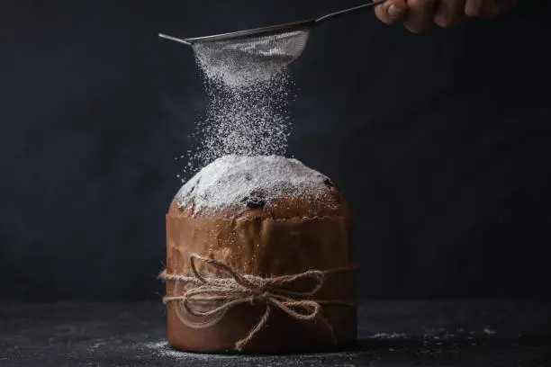 Traditional Italian Christmas cake Panettone tied with a jute thread on the black background. Pastry Chef sprinkles powdered sugar through a sieve on the fruit Easter cake with raisins.