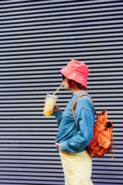istock Stylish girl in bright clothes and bucket hat drinking sugar flavored tapioca bubble tea while walking near gray striped urban wall. Portrait of fashionable hipster girl. Street fashion. Copy space. 1466738520