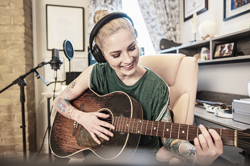 Female Musician practising music on acoustic guitar. She is dressed in casual clothes with sleeve tattoo on both arms.  Interior of home recording studio.