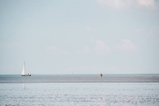 in the distance a person is walking through the Wadden Sea at low tide. A sailboat sails slowly on the horizon