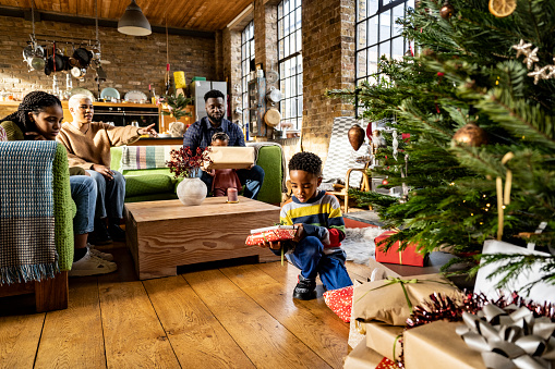 Young Black siblings enjoying holiday with parents and older sister, both excited to learn what is under wraps beneath decorated fir tree in loft apartment.