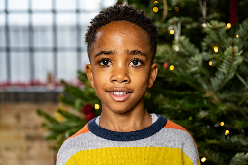 Head and shoulders view of shorthaired boy in striped sweater with round collar, standing in front of decorated Christmas tree and smiling at camera.