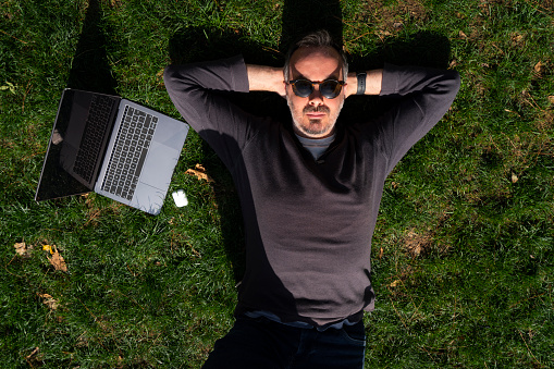 A mature man lying outside on the grass alongside a laptop and smartphone