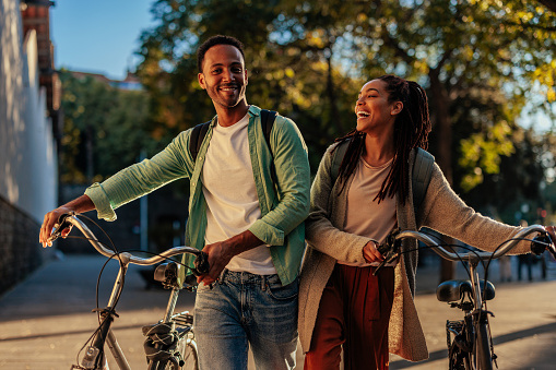 A young smiling couple is walking with their bicycles outside in the city on a sunny day.