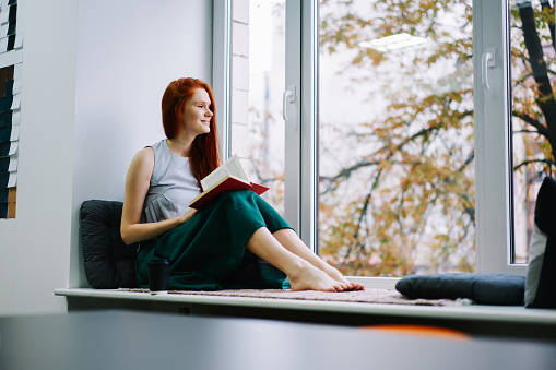 Full body barefoot young lady looking at window while sitting on comfortable windowsill with open novel while wearing casual outfit