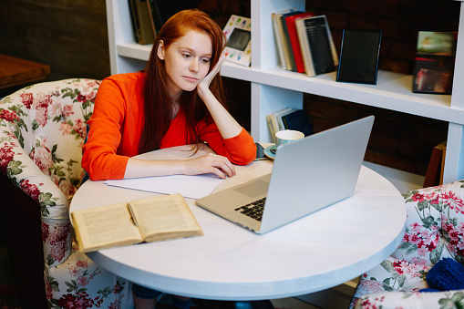 Young remote worker using computer and papers at table in cafe while sitting in armchair with mug and book and wearing casual clothes