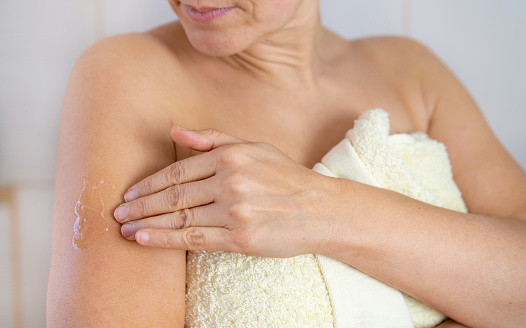 Close up of a woman wrapped in a towel after a shower as she applies an estrogen cream onto her arm, rubbing it in with her fingers