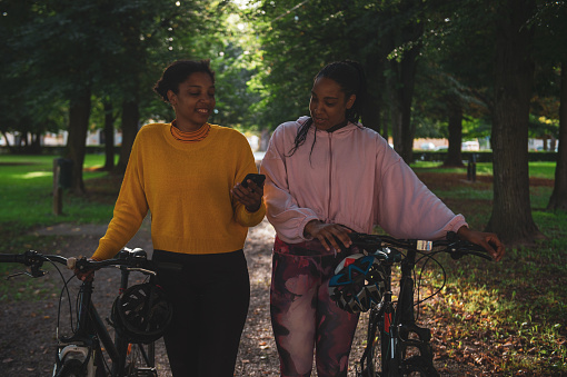 African American female athletes looking at map over smart phone and walking with bicycles in park against trees